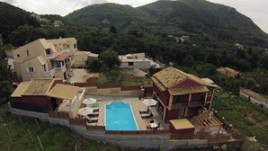 A residential area with a swimming pool, multiple houses, and a mountainous backdrop. - Villa Aris - Holiday & relaxation (Corfu - Kerkyra)
