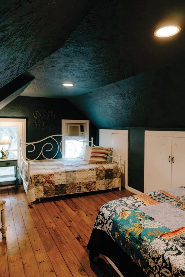 A bedroom with a sloped ceiling, a bed with a floral bedspread, a nightstand with a lamp, and a window with a view of the outdoors.