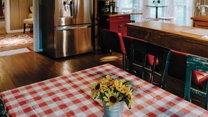 A kitchen with a stainless steel refrigerator, a wooden dining table with a red and white checkered tablecloth, and a vase of sunflowers.
