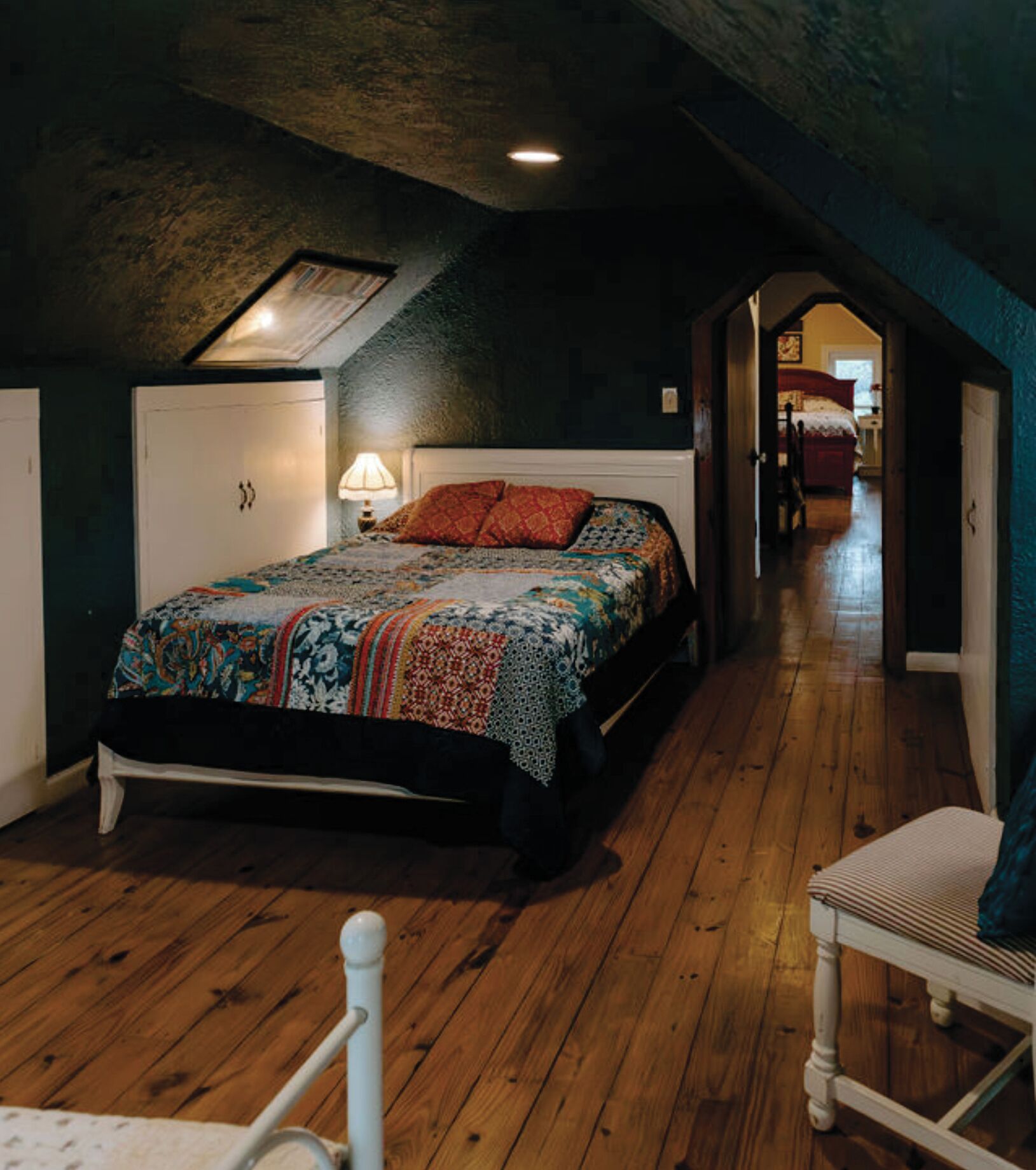 Ein Schlafzimmer mit einem großen Bett, einem Nachttisch, einer Lampe und einem Sessel. Ein Holzboden und eine Tür zu einem anderen Raum sind zu sehen.