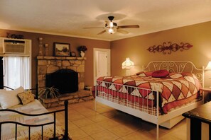 Une chambre avec une cheminée en pierre, un lit recouvert d’une couverture à motifs, un ventilateur de plafond et une table de chevet avec une lampe.
