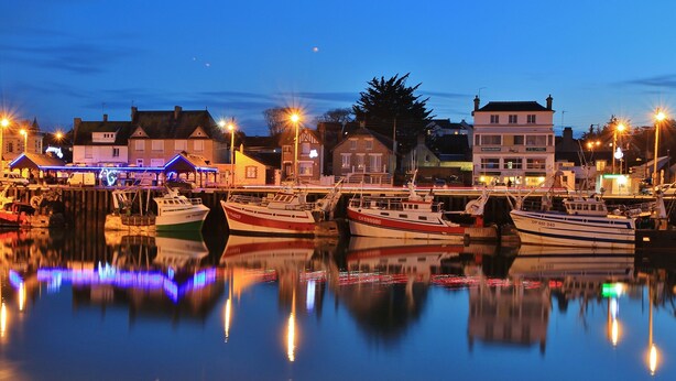 A harbor at dusk with boats docked, buildings with illuminated signs, and a calm water reflecting lights. - The wind rose 8 people (Grandcamp-Maisy)