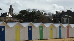 A row of beach huts with a church and houses in the background. - The wind rose 8 people (Grandcamp-Maisy)