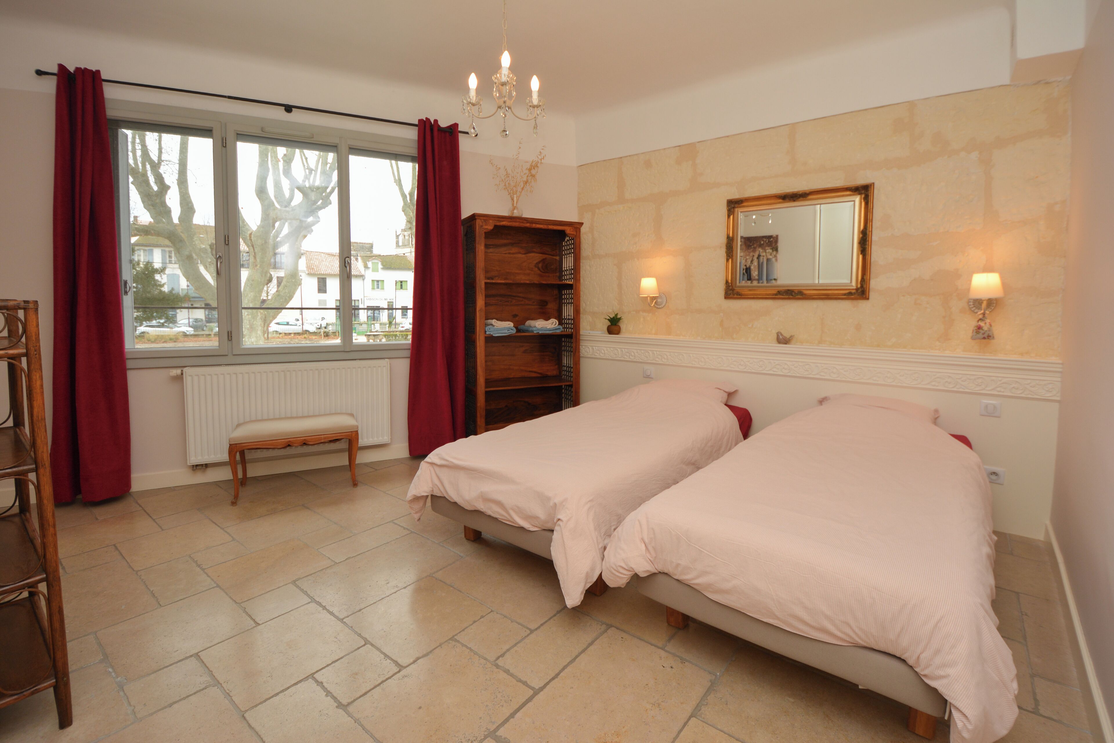 Une chambre avec deux lits, un banc en bois, un radiateur, une fenêtre avec des rideaux, un lustre, un miroir et une lampe fixée au mur.