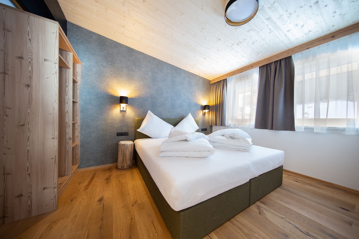 Una habitación de hotel moderna con suelo de madera, una cama grande con ropa blanca, dos luces fijadas a la pared y un ventanal con cortinas.