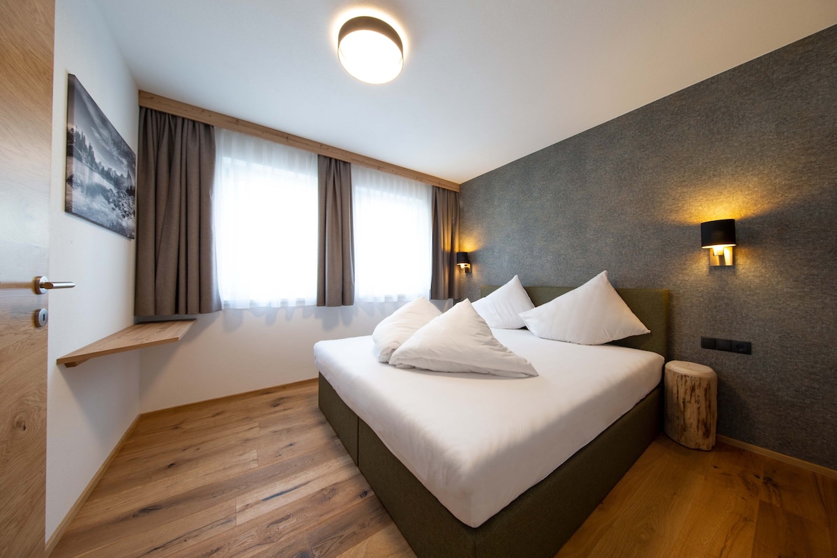 Un dormitorio moderno con una cama grande, suelo de madera, una ventana con cortinas y una lámpara fijada a la pared.