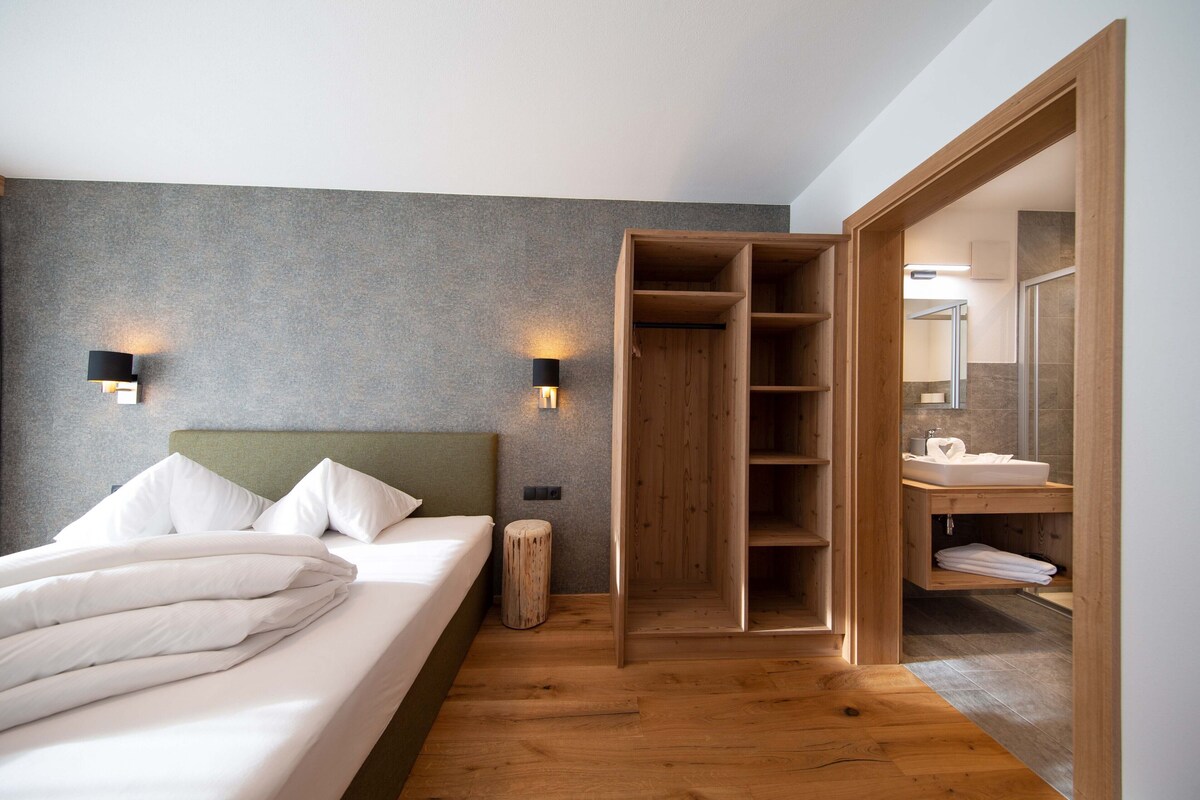 Un dormitorio con cama, lámparas de noche, un armario de madera y un baño con lavabo y espejo.