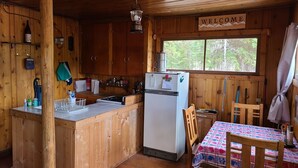 Una cabaña de madera con área de cocina, que incluye estufa, pileta y refrigerador. Se ve un cartel con la palabra 'WELCOME' en la pared, y hay una ventana con vista a árboles.
