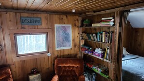 Interior acogedor de una cabaña con paredes de madera, una estantería llena de libros, una silla, una cama y un cartel que dice 'EAT SLEEP FISH'.