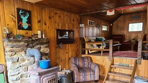 Interior rústico de una cabaña con chimenea de piedra, una cama litera de madera, un sillón estampado y un televisor montado.