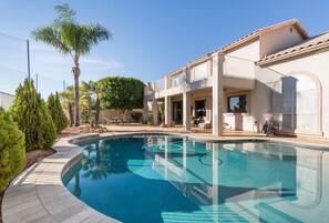 A swimming pool with a tiled deck, surrounded by a house with a balcony and a covered patio area.
