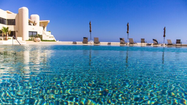 A clear pool with lounge chairs and umbrellas, a modern building with balconies, and a clear blue sky. - CABO Stunner~Ocean View Beach Condo with Gorgeous Sunsets Sleep 6+ (Cabo San Lucas)