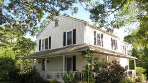 A two-story house with a porch, surrounded by lush greenery and flowering plants. - Charming Edgartown home with wrap around porch (Edgartown)