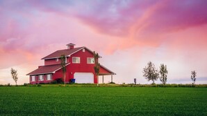 A red house with a garage, surrounded by a green field and trees, under a colorful sky. - Secluded 6 acre home near the Henry's Fork River. (Ashton)