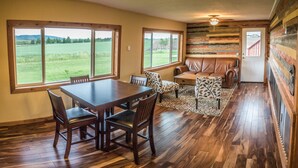 A dining area with a wooden table and chairs, a bench, and a view of a green field through large windows. - Secluded 6 acre home near the Henry's Fork River. (Ashton)