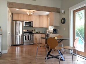 A modern kitchen with stainless steel appliances, wooden cabinets, and a dining area with a glass table and metal chairs. - Cottage in Historic Irvington neighborhood near Convention Center and shops! (Portland)