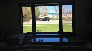 A living room with a large window overlooking a parking area and a grassy field. - BeachFrontRental-Tennis court&playground directly across the street!DogsWelcome (Elk Rapids)