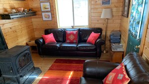 A cozy living room with a leather sofa, a red and orange rug, a wood-burning stove, and wooden walls. - Pristine Cabin near Craig...  Fish, or just Relax (Cascade)