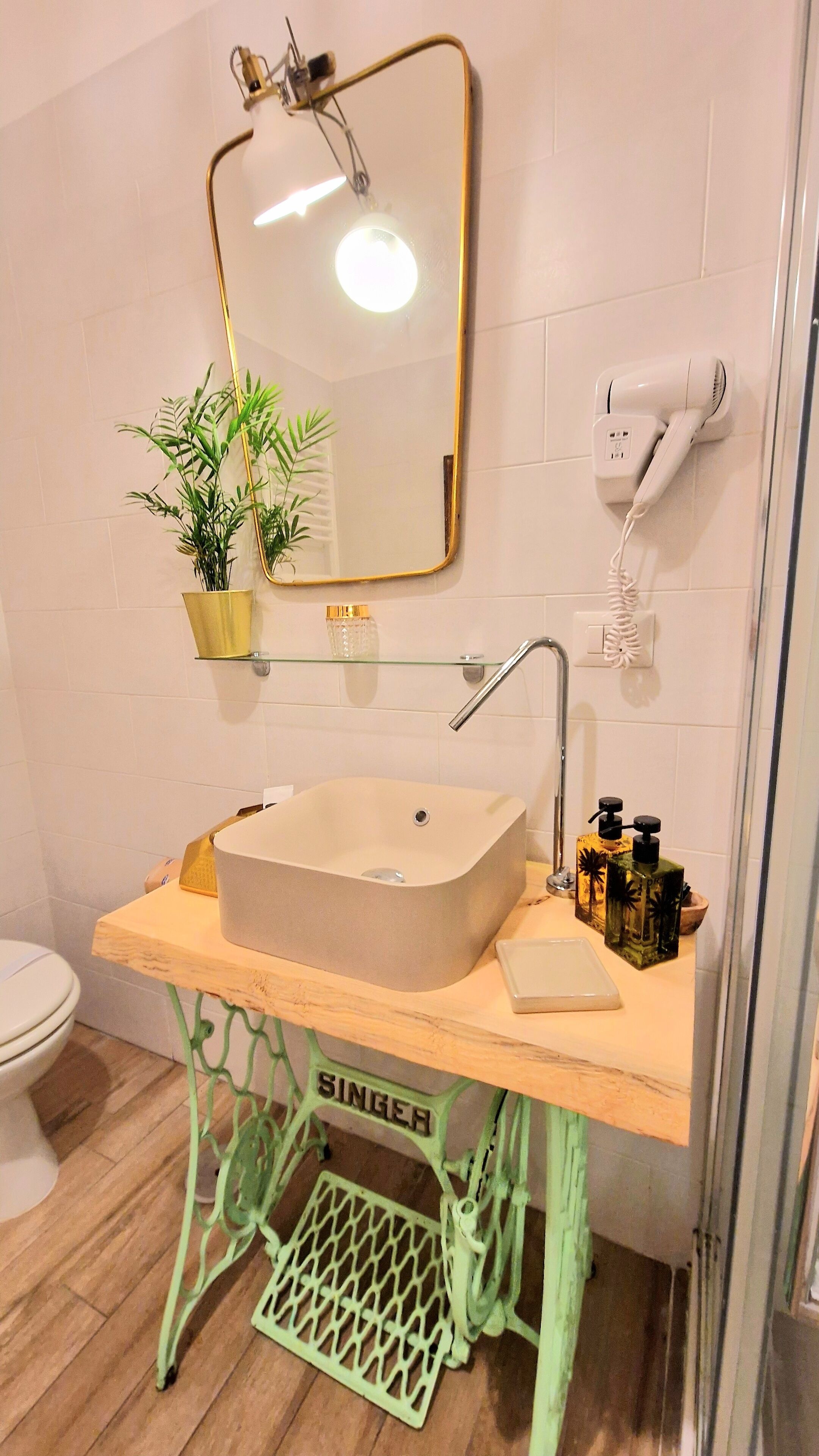 A bathroom with a beige sink, a mirror with a gold frame, a hairdryer, a potted plant, and a green metal stool.