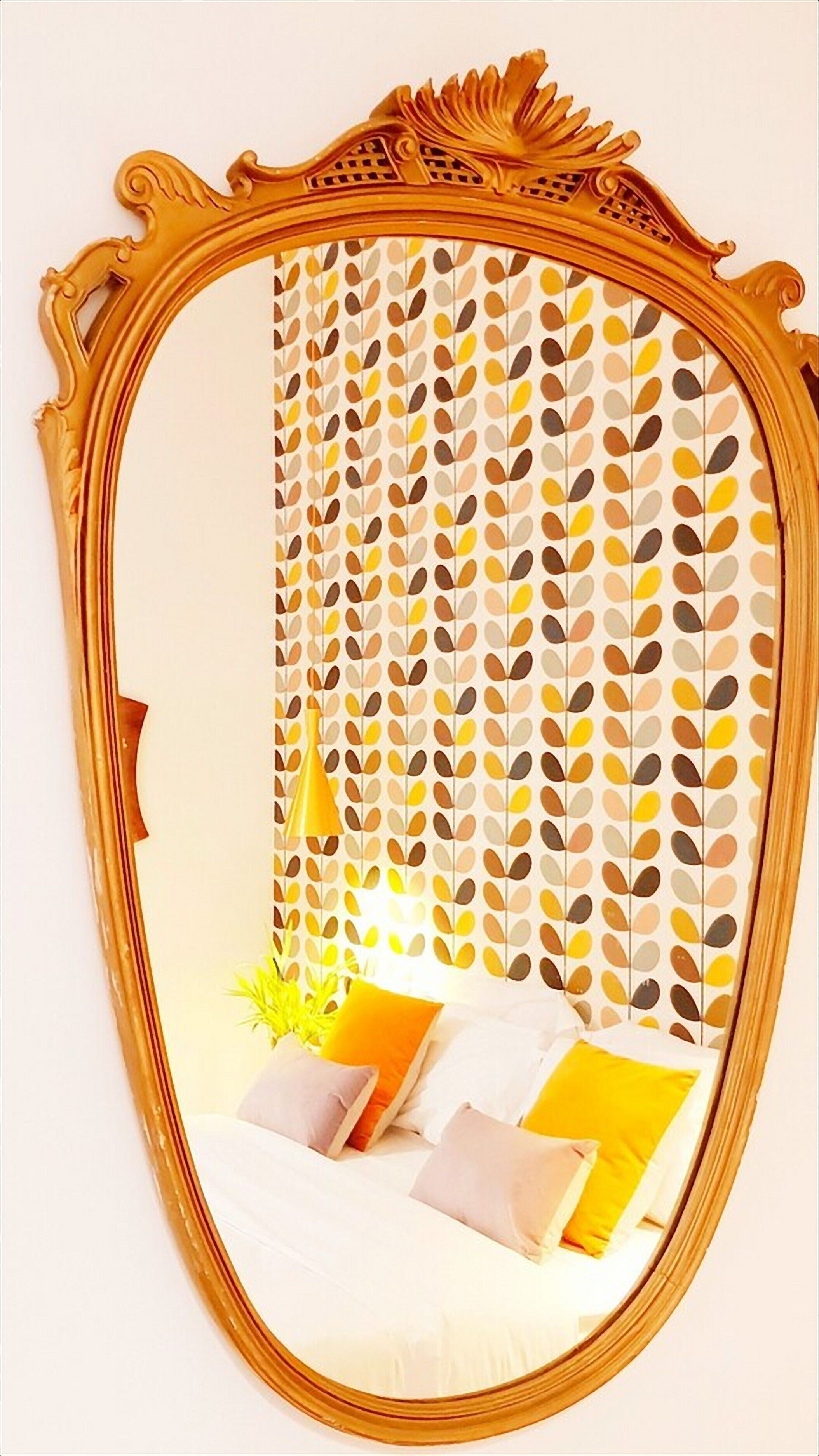 An oval mirror with a gold frame reflecting a bed with white bedding and multiple pillows, including a yellow one.