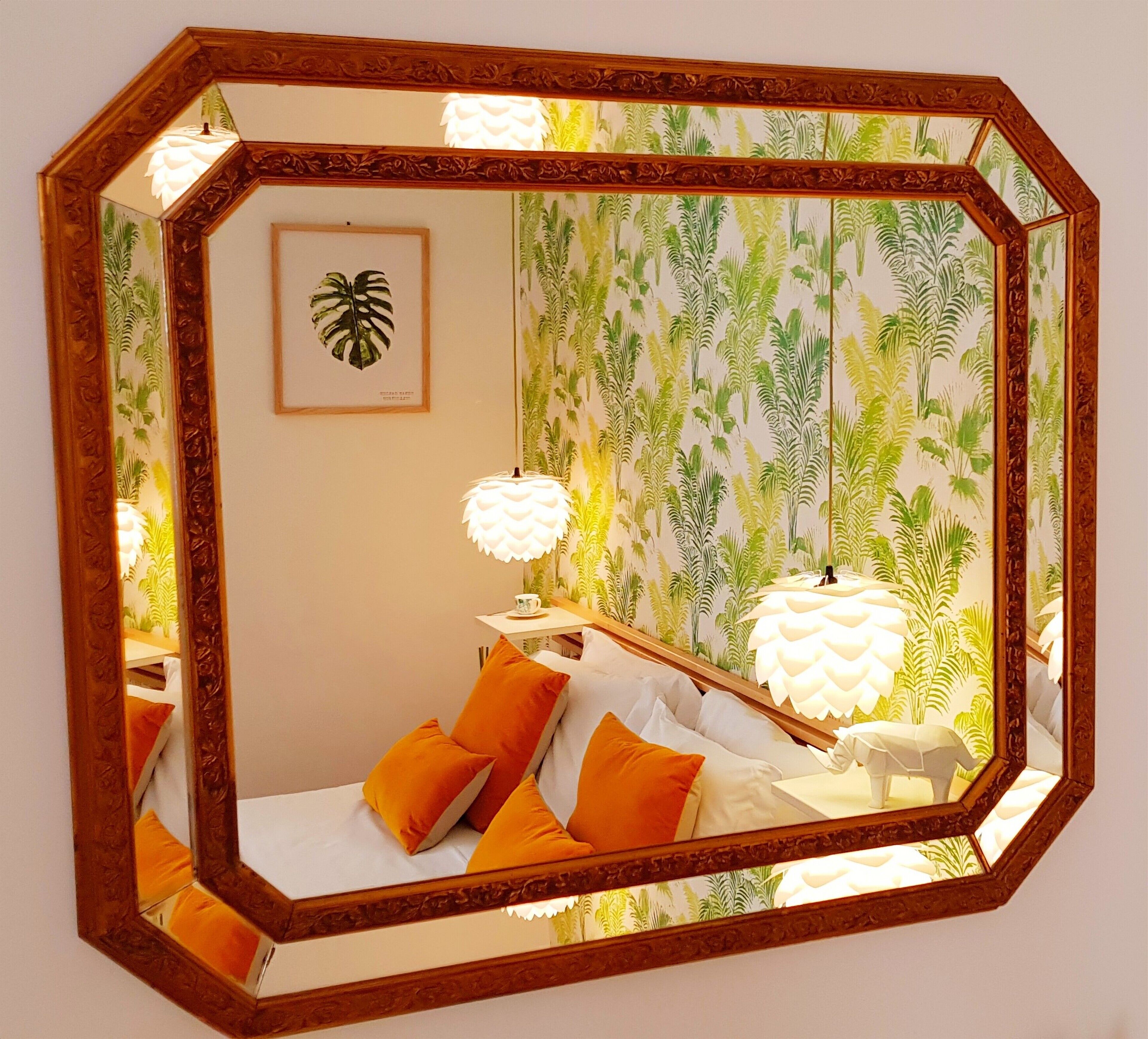 A bedroom with a large octagonal mirror, a bed with orange pillows, a nightstand with a lamp, and a tropical wallpaper.