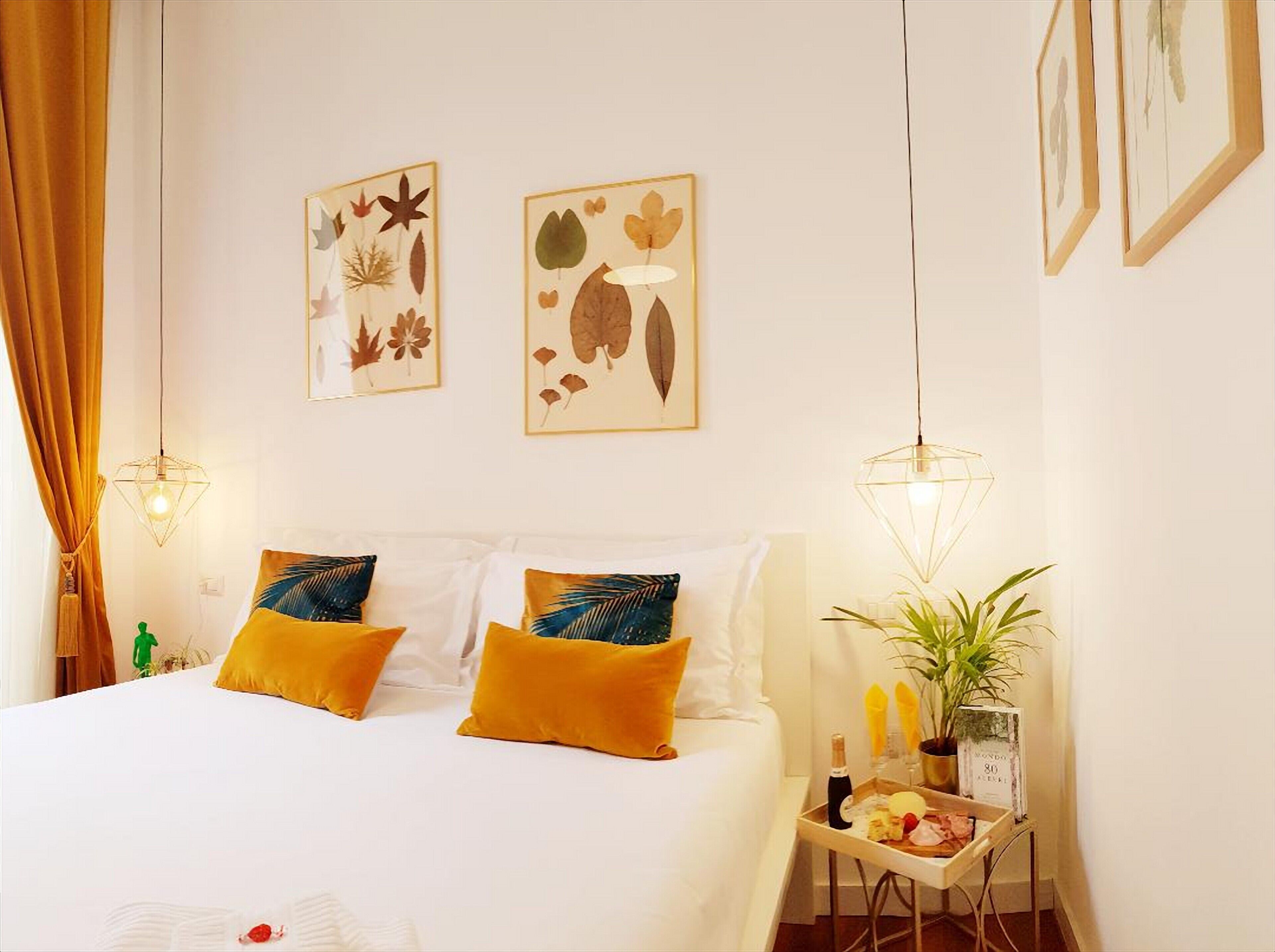 A bedroom with a bed, two wall-mounted lamps, a nightstand with a plant and a tray of food, and two framed botanical prints on the wall.