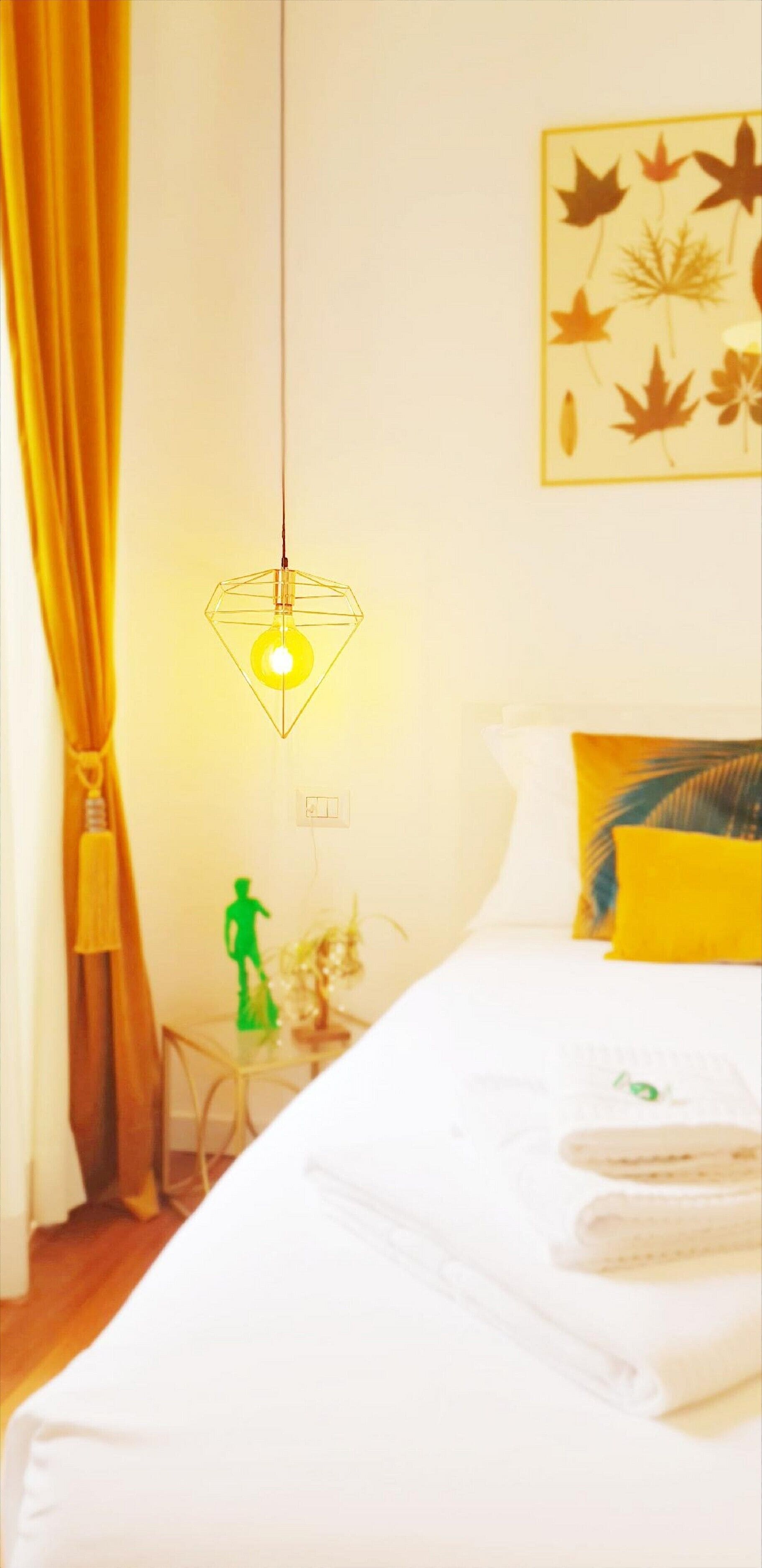 Een hotelkamer met een bed, een hanglamp, een muur met een bladpatroon en een tafeltje met een groene figuur en een plant.