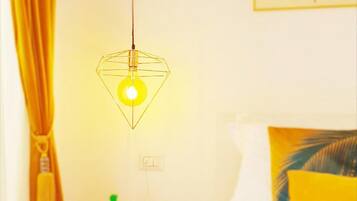 Een hotelkamer met een bed, een hanglamp, een muur met een bladpatroon en een tafeltje met een groene figuur en een plant.