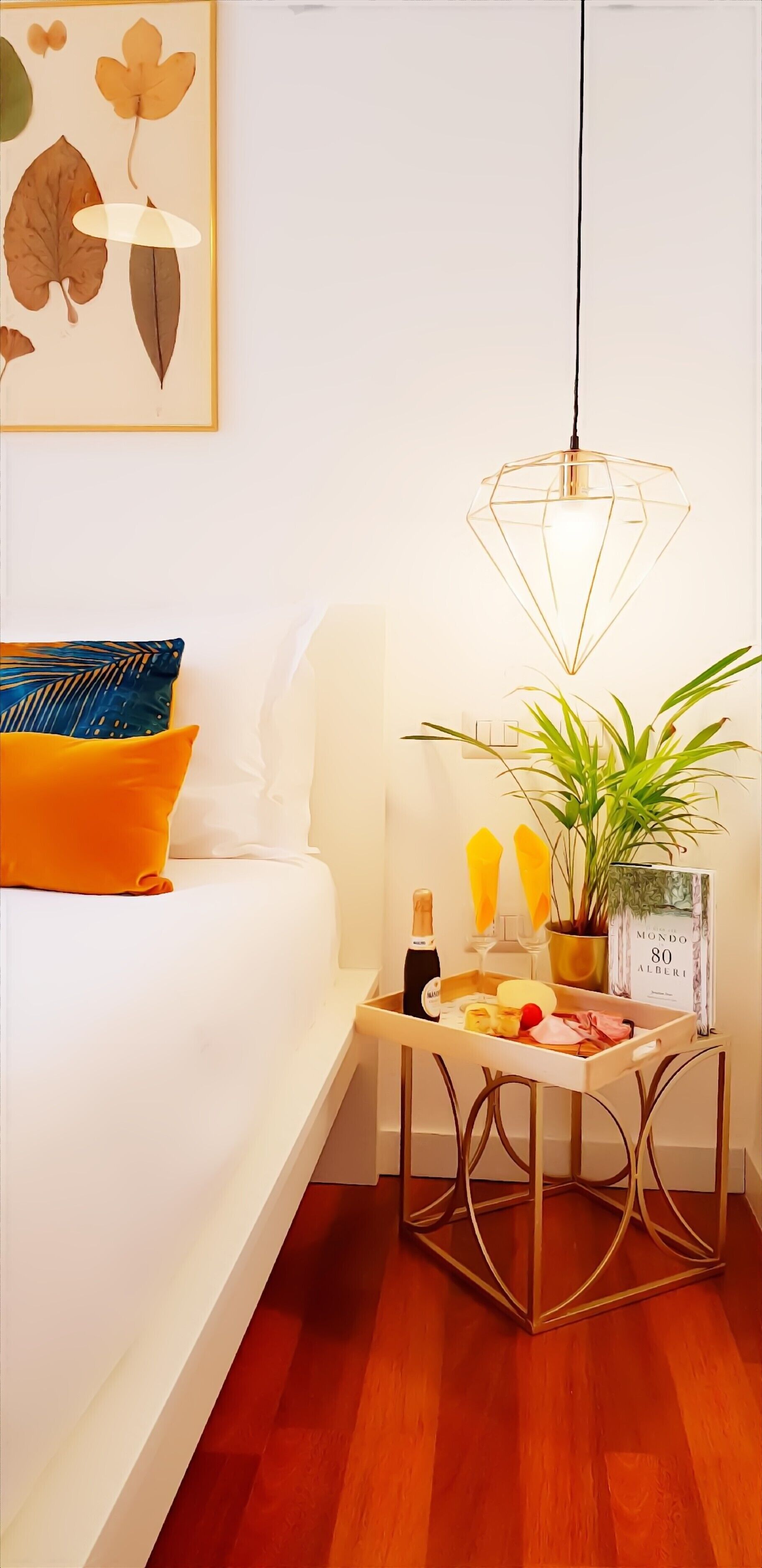 Une chambre à coucher avec un lit, une table de chevet sur laquelle se trouvent une bouteille, des verres et une assiette de nourriture, un luminaire suspendu et une plante.