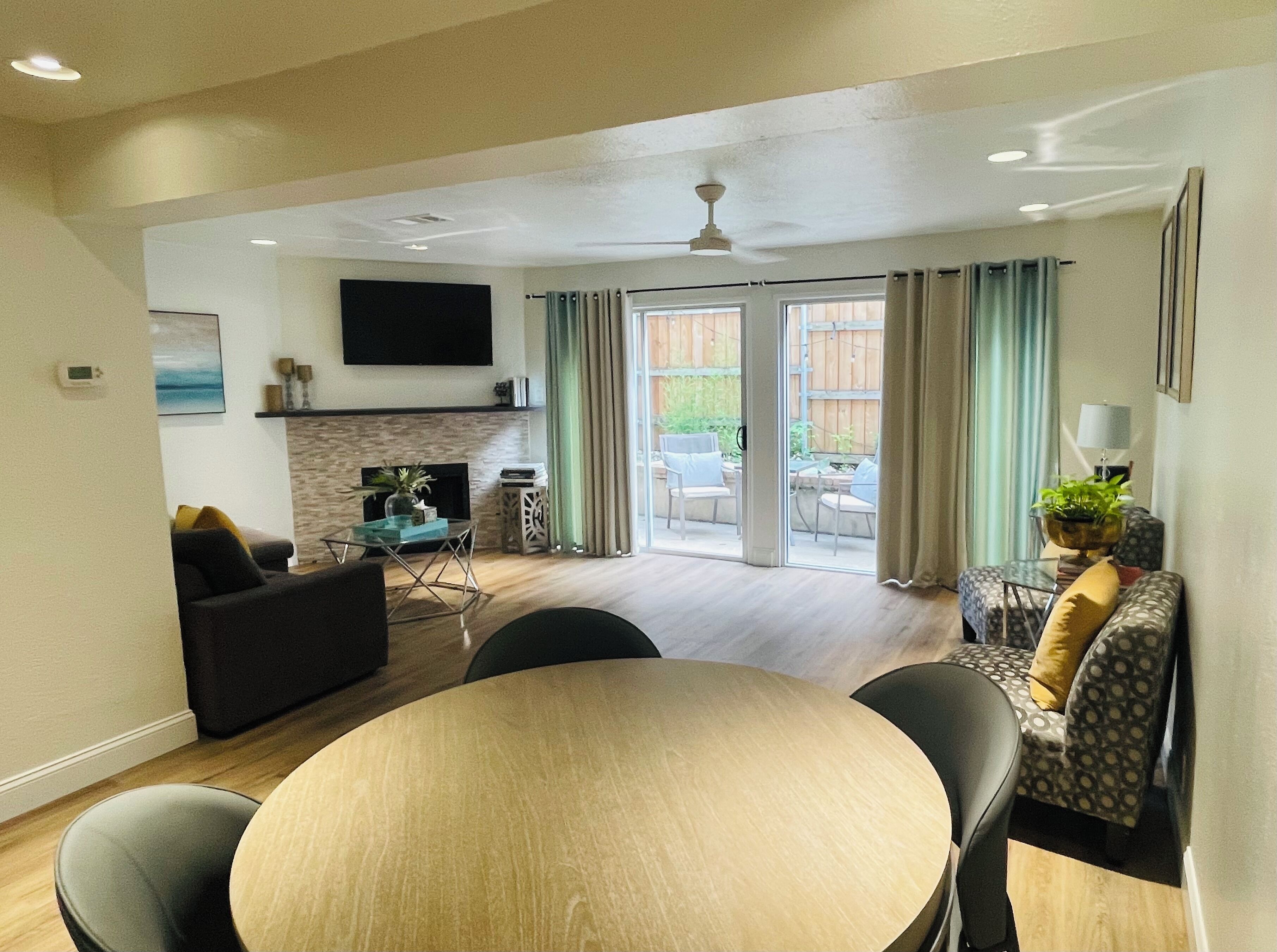 A modern living room with a fireplace, a dining area with a round table, and a large window with curtains.