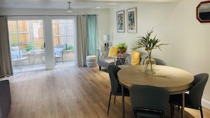Ruime kamer met houten vloer, een ronde eettafel met stoelen, een plafondventilator en een schuifdeur van glas die leidt naar een terras met een stoel en een tafel.