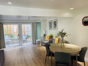 A spacious room with a wooden floor, a round dining table with chairs, a ceiling fan, and a sliding glass door leading to an outdoor area with a chair and a table.