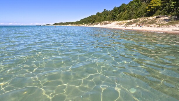 Ein Strand mit klarem, flachen Wasser, einem Sandstrand und einem bewaldeten Hügelhang.