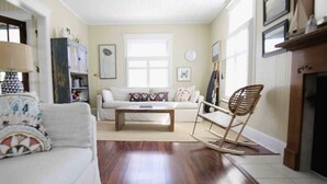 Ein Wohnzimmer mit einem weißen Sofa, einem Holztisch und einem Schaukelstuhl. Rechts befindet sich ein Kamin und im Hintergrund steht ein Bücherregal.