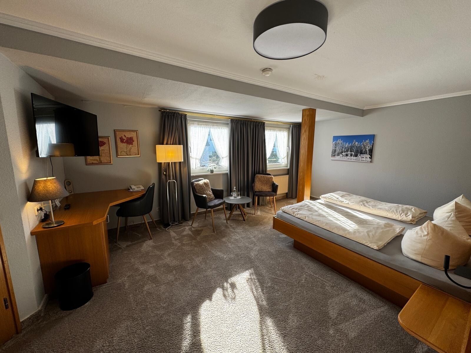 A hotel room with a bed, desk, chairs, a TV, and a window with curtains.