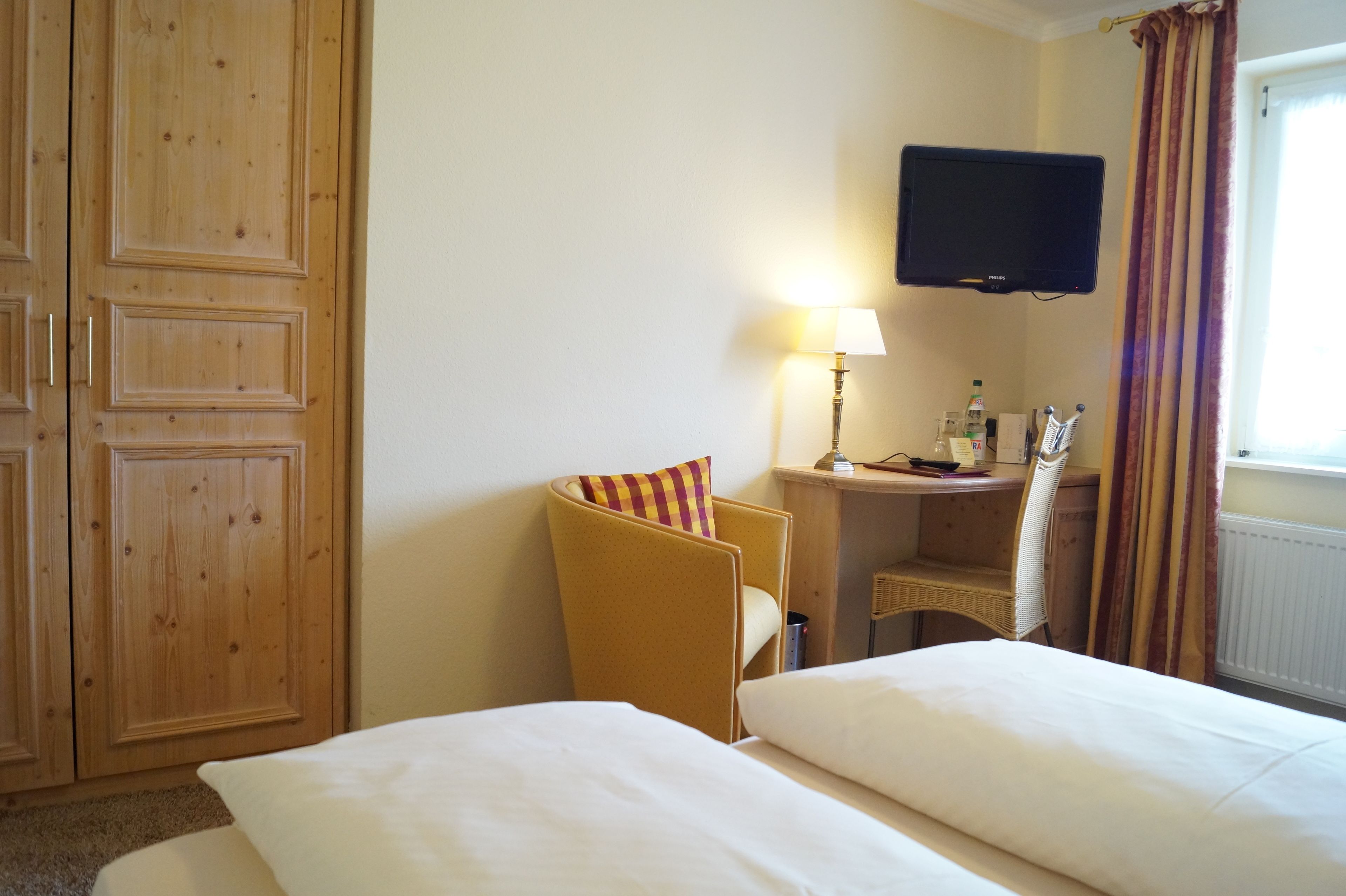 Ein Hotelzimmer mit zwei Betten, einem Holzschrank, einem Schreibtisch mit Lampe, einem Fernseher und einem Fenster mit Vorhängen.