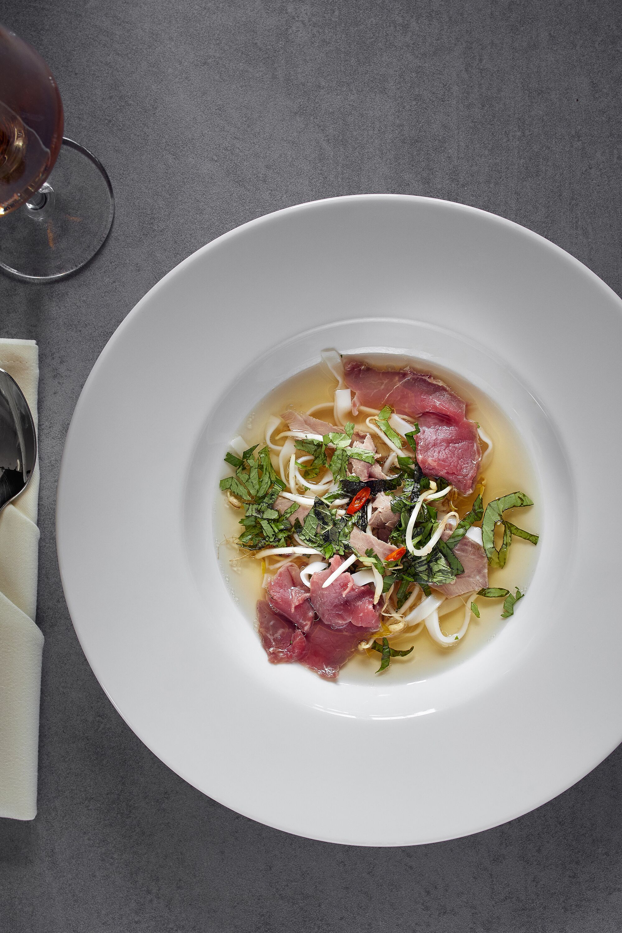 Uma tigela branca de sopa com macarrão, carne e ervas sobre uma superfície cinzenta.