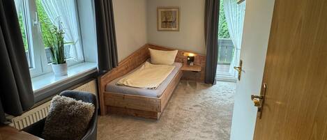 Un dormitorio pequeño con una cama individual, una mesita de noche, un radiador, una ventana con cortinas y una puerta.