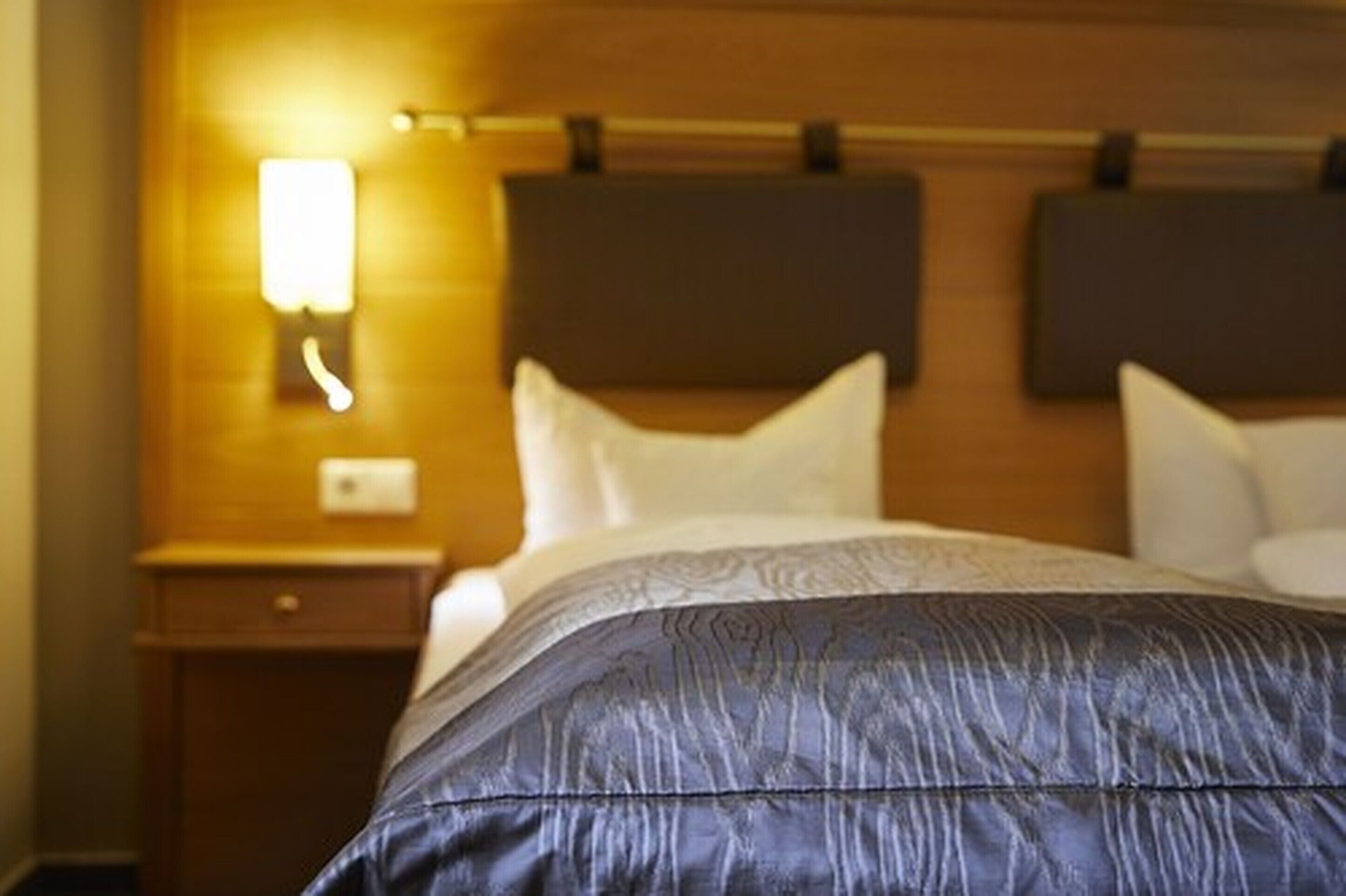 A hotel room with a neatly made bed, a wooden headboard, and a nightstand with a lamp.