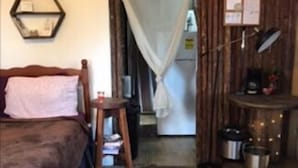 Una habitación rústica con una cama de madera, un taburete pequeño, un refrigerador y una estantería con ollas.