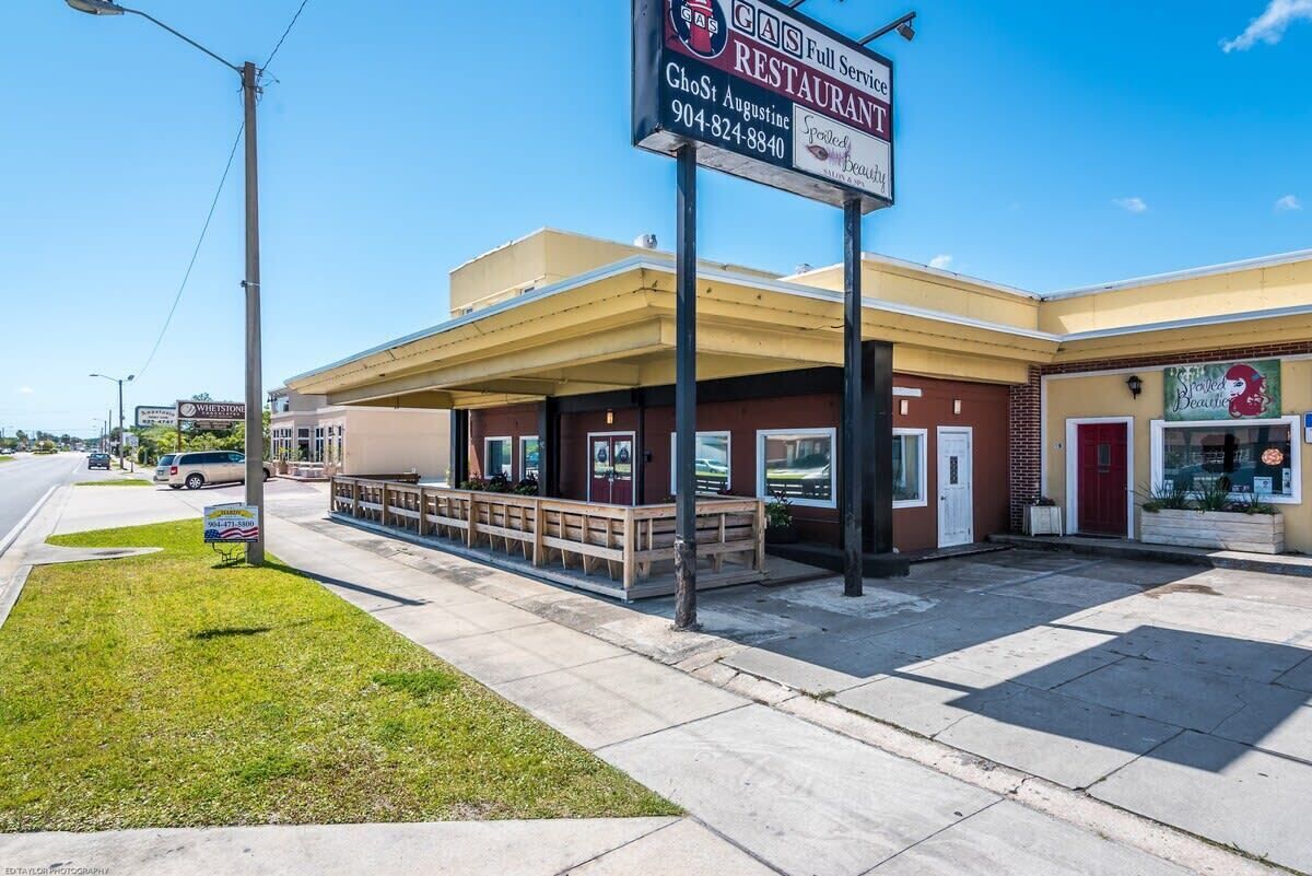 Un restaurant avec une terrasse, un panneau indiquant une station-service et une devanture de magasin avec une porte rouge.