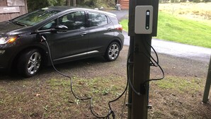 Een auto geparkeerd bij een laadstation voor elektrische voertuigen, met een hutje genaamd "Cabin Wildwood" op de achtergrond.