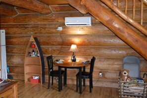 A wooden cabin interior with a dining table, chairs, a lamp, a teddy bear, and a small shelf with bottles. - Cabin In The Wildwood (Chehalis)