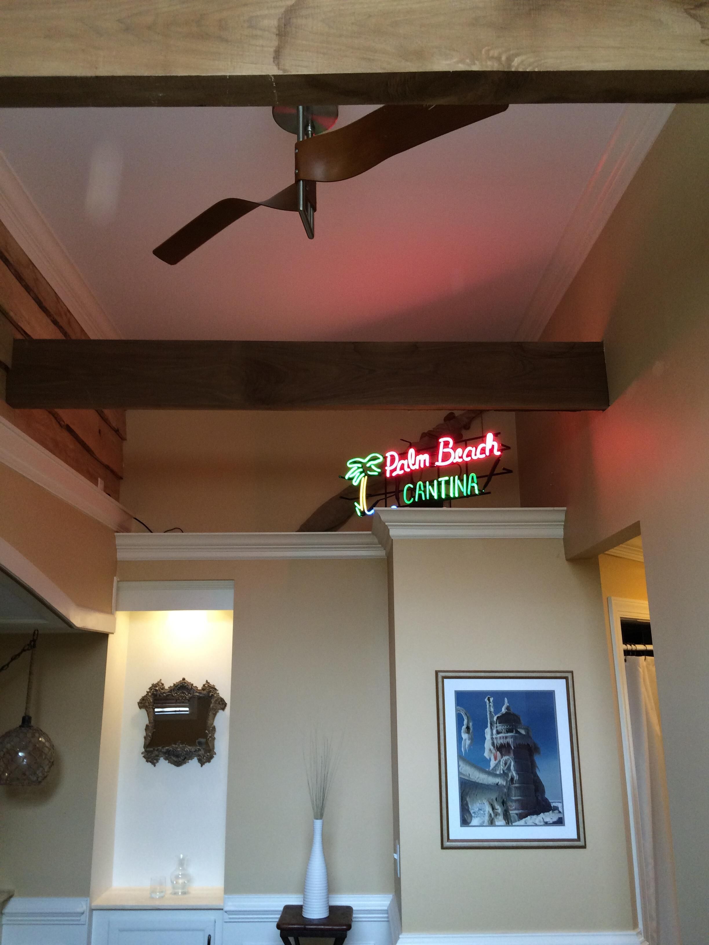 Una habitación con ventilador de techo, un letrero de neón que dice "Palm Beach Cantina", un cuadro en la pared y un florero sobre una mesita.