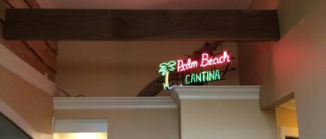 Una habitación con ventilador de techo, un letrero de neón que dice "Palm Beach Cantina", un cuadro en la pared y un florero sobre una mesita.