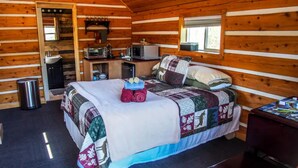 Ein Blockhausszimmer mit Bett, einer kleinen Küchenzeile, einem Fenster und einem Mülleimer.