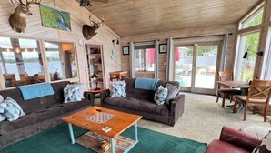 A cozy cabin interior with a wooden ceiling, a ceiling fan, a fireplace, and a large window overlooking a scenic view. - Cottage on a Lake.  Near Pictured Rocks/ Munising (Manistique)