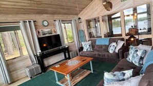 A cozy cabin interior with a wooden ceiling, a fireplace, a television, and a large window overlooking a lake. - Cottage on a Lake.  Near Pictured Rocks/ Munising (Manistique)