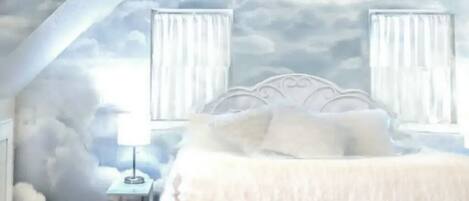 A bedroom with a sloped ceiling, a bed with white bedding, two bedside tables with lamps, and a wall mural of clouds.