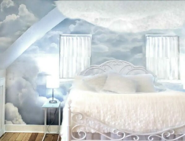 A bedroom with a sloped ceiling, a bed with white bedding, two bedside tables with lamps, and a wall mural of clouds.
